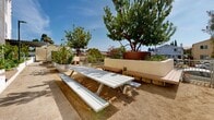 Located at 4340 44th Street in San Diego's City Heights neighborhood, this outdoor community space serves as a vibrant garden corridor nestled alongside a modern multi-story residential building. The space is thoughtfully designed to encourage community gathering and urban agriculture, stretching as a long, well-organized outdoor passageway filled with greenery and gathering amenities.

Raised garden beds constructed from whitewashed concrete block line the pathway and overflow with a diverse array of plants, including fruit trees, grapevines, ornamental grasses, herbs, and vegetables. Large galvanized metal containers and additional planter boxes punctuate the space, offering further growing areas and visual interest. Mature trees provide natural shade and contribute to a lush, park-like atmosphere under the sunny San Diego sky.

The hardscaped walkway is complemented by warm wood-toned composite benches integrated into the planter walls, offering casual seating throughout. A picnic table with bench seating provides a dedicated gathering spot, ideal for communal meals or outdoor activities. Street-level lighting fixtures ensure the space remains accessible and welcoming at all hours.

The adjacent building features a contemporary façade with stucco cladding, bold accent colors, and large windows, adding architectural character to the setting. Overall, this outdoor space combines functional urban gardening with comfortable communal amenities, creating an engaging, green retreat within the city.