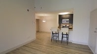 This single-floor space showcases an open-concept layout that seamlessly connects the living area to the kitchen. Light-toned hardwood flooring extends throughout, lending warmth and visual continuity to the interior. The soaring ceilings amplify the sense of space, creating an airy, expansive atmosphere accentuated by clean white walls.

The kitchen is a standout feature, equipped with dark espresso cabinetry that contrasts elegantly against a stone tile backsplash. Stainless steel appliances, including a range and over-the-range microwave, add a modern, functional touch. A peninsula-style breakfast bar with a light stone countertop extends into the living area, accompanied by two bar-height chairs with dark frames, making it an ideal spot for casual dining or entertaining.

A pendant light fixture hangs gracefully above the living area, providing focused ambient lighting that complements the recessed ceiling light in the kitchen. A hallway visible in the background connects additional areas of the space. Overall, the design balances contemporary aesthetics with practical functionality, creating a cohesive and inviting environment.