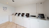 This single-floor shared laundry facility is designed for practical, high-capacity use. The layout is straightforward and efficient, with a row of top-loading, coin-operated washing machines lining one wall atop a continuous white cabinet base. Opposite the washers, two sets of stacked coin-operated dryers provide ample drying capacity to match the volume of the washing stations. The flooring throughout consists of large-format light beige tiles arranged in a diagonal pattern, contributing to a clean, open feel underfoot.

The walls are finished in a neutral white, keeping the space bright and easy to maintain. Recessed lighting in the textured ceiling provides even illumination across the room. Natural light enters through a glazed entry door flanked by sidelight windows, adding a welcoming quality to the utilitarian space. A waste bin is conveniently positioned near the dryers, and informational signage is posted on the walls for user guidance.

The entry door features a standard door closer for controlled access, and the overall layout provides adequate open floor space for residents to move comfortably while managing laundry tasks. The space is functional, well-lit, and clearly maintained for shared residential use.