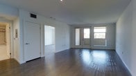 This clean and contemporary single-floor space is designed with a minimalist aesthetic that emphasizes openness and functionality. The main living area is expansive and airy, showcasing rich, dark hardwood floors that extend throughout and contrast elegantly with the crisp white walls and ceiling. A set of large glass-paneled French doors anchors one wall, flooding the interior with natural light while offering direct access to an outdoor area and views of the surrounding neighborhood skyline. The open layout allows for flexible furniture arrangement, making the space highly adaptable to a variety of uses. An adjacent room, visible through a wide doorway, provides additional versatile space with the same bright, neutral palette and hardwood flooring, maintaining a consistent and cohesive design language throughout. Recessed lighting in the ceiling supplements the natural light, ensuring the interior remains well-lit at all hours. The overall ambiance is calm, modern, and inviting — a blank canvas that combines practical design with understated elegance. Ceiling-mounted HVAC vents are thoughtfully integrated, contributing to a seamless and uncluttered finish across the entire space.