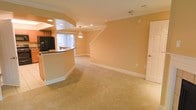 This two-story home offers a welcoming and well-appointed living environment with a thoughtful layout spread across two floors. The main level showcases an open-concept design where the kitchen flows seamlessly into the living area, creating a spacious and functional gathering space. The kitchen features a curved breakfast bar with dark countertops, recessed cabinetry, and a black refrigerator, while a large window dressed with vertical blinds allows natural light to fill the space. A decorative pendant light hangs above the kitchen bar area, adding a refined touch to the transition between spaces. The living area is finished with plush, neutral-toned carpeting that lends warmth underfoot, while the walls are painted in a soft, golden hue that contributes to the overall inviting atmosphere. Crown molding lines the ceilings, adding an elegant architectural detail throughout. A staircase with a clean white balustrade leads to the upper floor, providing access to the home's additional living spaces. Recessed lighting is thoughtfully placed across the ceiling, ensuring even, ambient illumination. White baseboards and panel doors complete the classic interior aesthetic, giving the home a timeless and polished character overall.