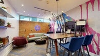 This dynamic single-room studio is a visually striking creative space that masterfully combines industrial character with contemporary design. The centerpiece of the room is a dramatic large-scale mural featuring bold geometric shapes and vivid splashes of pink, blue, and black, commanding attention and setting an energetic tone throughout. Contrasting beautifully, an exposed brick wall adds raw, industrial texture alongside a neon sign that contributes a playful, urban edge.

The space is thoughtfully zoned for multiple uses. A high-top wooden conference table surrounded by sleek navy blue chairs anchors the collaborative work area, equipped with professional podcasting and recording equipment including microphones, monitors, and ring lighting — making it well-suited for content creation and media production. A lounge area with a sage green sofa, accent chairs, and a sculptural coffee table provides a comfortable setting for casual conversation or relaxation.

Acoustic wall panels in varying shades of grey and navy line one wall, enhancing sound quality within the space. A chevron-patterned carpet with blue accents flows across the floor, while a multi-arm Edison bulb chandelier overhead adds a modern industrial lighting element. A glass-panel door connects to a bright corridor, allowing natural light to filter in while maintaining privacy.