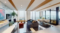 This elegantly designed clubroom occupies a single floor high above the city, offering panoramic skyline views through expansive floor-to-ceiling glass walls that flood the space with natural light. The open-plan main lounge features a striking high-gloss checkerboard floor, exposed natural wood ceiling beams, and recessed lighting, creating a refined yet welcoming atmosphere. A plush sectional sofa anchors the seating area, complemented by an adjacent dining setup with sleek gold-accented chairs and a round table surrounded by views on all sides.

A secondary lounge area showcases warm hardwood flooring, spindle-leg armchairs, and a curated burl wood coffee table, giving it a more intimate, residential character. Bold artwork displayed against a graphic black-and-white patterned accent wall adds a gallery-like quality to yet another sitting area.

The space also includes two kitchenette areas — one featuring vibrant teal cabinetry, warm stone countertops, and a pass-through window, and another with sleek dark stone surfaces and light wood cabinetry. A graceful arched brick entryway connects a wide corridor lined with private rooms and additional amenity spaces, making this clubroom a versatile and visually compelling destination.