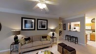 Located at 8400 Brass Mill Ln in Raleigh, NC, this single-floor residence offers a well-designed layout with a cohesive, neutral color palette throughout. The living area serves as the heart of the home, featuring plush carpeting, a ceiling fan with integrated lighting, and tasteful wall art that adds a refined touch to the space. A comfortable sectional sofa anchors the room, complemented by woven ottoman seating and warm accent lighting from a classic table lamp, creating an inviting and relaxed atmosphere. Crown molding along the upper walls adds an elegant architectural detail. An open pass-through connects the living area to the kitchen, enhancing the sense of openness and allowing natural sightlines across the space. The kitchen features light countertops, bar-height seating, and ample cabinetry, making it both functional and accessible. The open-concept design encourages easy flow between the living and dining areas, with a glimpse of additional rooms beyond, suggesting a thoughtfully organized floor plan. The overall ambiance is warm and comfortable, with a neutral tone palette of beige, gray, and dark accents that lends a modern yet approachable character to this Raleigh residence.