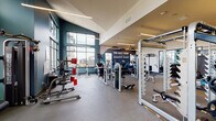 Located at 22004 E Quincy Ave in Aurora, Colorado, this fitness center offers a thoughtfully designed, single-floor workout facility that blends functionality with modern aesthetic appeal. The space features soaring ceilings that enhance the sense of openness, while large, floor-to-ceiling windows along the teal-accented walls flood the interior with natural light and provide views of the surrounding neighborhood.

The facility is divided into distinct workout zones. The cardio area houses a row of StairMaster steppers and elliptical machines, positioned in front of a striking motivational wall mural depicting a mountain landscape with the phrase "The Best View Comes After the Hardest Climb." This feature wall serves as both a design centerpiece and an inspirational focal point.

The strength training section is generously outfitted with a variety of cable machines, selectorized weight machines, a squat rack, benches, and free weights including a dumbbell rack. Equipment from brands such as TechnoGym and Nautilus is prominently featured throughout.

Linear pendant lighting runs along the ceiling, complemented by ceiling fans that maintain airflow and comfort. Full-length mirrors are strategically placed to support proper form during workouts. A set of glass-panel double doors provides easy access to the facility, reinforcing the open, welcoming atmosphere of this well-appointed fitness space.