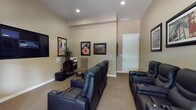 This single-floor home theater room is thoughtfully designed to deliver an immersive cinematic experience. The space is finished with warm, neutral-toned walls and plush wall-to-wall carpeting, creating a comfortable and acoustically favorable environment. Multiple rows of black leather reclining theater seats with built-in cup holders are arranged to provide clear sightlines to the large wall-mounted flat-screen television, which serves as the room's focal point. Recessed ceiling lighting provides ambient illumination, complementing the overall mood of the space.

The room is decorated with framed classic movie posters and cinema-themed artwork, reinforcing its dedicated entertainment purpose. A media console and subwoofer are positioned near the screen, suggesting a full audio-visual setup. A natural wood-trimmed window with horizontal blinds along one wall allows for controlled natural light when desired, while a potted plant adds a touch of warmth to the otherwise purposeful décor. Additional accent chairs provide flexible seating options. A private door offers convenient access, completing this well-appointed and functional home theater space.