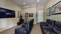 This single-floor home theater room is thoughtfully designed to deliver an immersive cinematic experience. The space is finished with warm, neutral-toned walls and plush wall-to-wall carpeting, creating a comfortable and acoustically favorable environment. Multiple rows of black leather reclining theater seats with built-in cup holders are arranged to provide clear sightlines to the large wall-mounted flat-screen television, which serves as the room's focal point. Recessed ceiling lighting provides ambient illumination, complementing the overall mood of the space.

The room is decorated with framed classic movie posters and cinema-themed artwork, reinforcing its dedicated entertainment purpose. A media console and subwoofer are positioned near the screen, suggesting a full audio-visual setup. A natural wood-trimmed window with horizontal blinds along one wall allows for controlled natural light when desired, while a potted plant adds a touch of warmth to the otherwise purposeful décor. Additional accent chairs provide flexible seating options. A private door offers convenient access, completing this well-appointed and functional home theater space.