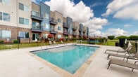 This outdoor amenity space centers around a generously sized rectangular swimming pool with clear, blue-tinted water and a shallow entry area, making it accessible and inviting. The pool deck is finished in light-toned concrete pavers, providing ample room for relaxation and circulation. Multiple lounge chairs are positioned around the perimeter, offering comfortable spots to enjoy the open sky and sunshine.

The pool area is enclosed by a black wrought iron security fence with a gated entry, ensuring safety and controlled access. Posted pool rules and safety equipment, including a life ring, are conveniently located near the entrance. A utility storage shed is situated along one side of the enclosure.

The surrounding residential buildings feature a contemporary design, combining brick bases with gray and blue panel siding, and each unit includes private balconies that overlook the pool and grounds below. Lush landscaping with flowering shrubs and manicured lawn areas adds color and greenery to the environment.

Beyond the fence line, open green space and a secondary recreational area — including what appears to be a volleyball or sport court — extend the outdoor experience. The broader setting features wide-open views under expansive skies, creating a bright and airy atmosphere throughout the entire outdoor amenity area.