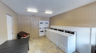 This single-floor community space is designed to serve residents with functional, well-maintained shared amenities. The laundry facility is a practical and organized room, equipped with multiple top-load washing machines and stacked washer-dryer units lined along the walls. The room features warm wood-paneled walls, a textured ceiling with flush fluorescent lighting, and durable stone-tile flooring in neutral tones. A storage closet and seating area with a wicker chair add to the room's utility and comfort.

The outdoor area centers around a generously sized swimming pool with clear blue water, surrounded by a wide, light-colored concrete deck. Sun loungers are positioned poolside, inviting relaxation in the open air. The pool area is enclosed by a wrought iron fence and framed by mature trees that provide natural shade and greenery. The surrounding multi-story residential buildings with brick and wood accents are visible in the background, reinforcing the community atmosphere. Under open skies, the outdoor space offers a bright and inviting environment ideal for leisure and recreation.