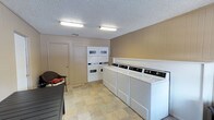This single-floor community space is designed to serve residents with functional, well-maintained shared amenities. The laundry facility is a practical and organized room, equipped with multiple top-load washing machines and stacked washer-dryer units lined along the walls. The room features warm wood-paneled walls, a textured ceiling with flush fluorescent lighting, and durable stone-tile flooring in neutral tones. A storage closet and seating area with a wicker chair add to the room's utility and comfort.

The outdoor area centers around a generously sized swimming pool with clear blue water, surrounded by a wide, light-colored concrete deck. Sun loungers are positioned poolside, inviting relaxation in the open air. The pool area is enclosed by a wrought iron fence and framed by mature trees that provide natural shade and greenery. The surrounding multi-story residential buildings with brick and wood accents are visible in the background, reinforcing the community atmosphere. Under open skies, the outdoor space offers a bright and inviting environment ideal for leisure and recreation.