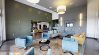 220 Lounge is a thoughtfully designed, single-floor communal space that blends modern aesthetics with a welcoming, social atmosphere. The open-concept layout flows seamlessly across several distinct zones, each serving a unique purpose while maintaining a cohesive design language throughout.

The main lounge area features soaring ceilings with large pendant light fixtures that cast a warm, inviting glow over a curated arrangement of soft blue accent chairs, a neutral sectional sofa, and a circular patterned area rug. Floor-to-ceiling windows allow abundant natural light to pour in, enhancing the airy, spacious feel of the room.

A bold olive-green shiplap accent wall creates a striking architectural focal point, visually separating the primary lounge from an adjacent seating nook furnished with mustard-yellow chairs and additional casual seating. This secondary zone offers a more intimate setting within the broader open floor plan.

Toward the rear of the space, a bar and counter service area provides a central hub for refreshments, complete with bar-height seating and sleek cabinetry. Display cases and gallery-style wall features add refined detail throughout.

The overall ambiance of 220 Lounge is contemporary and polished, combining clean lines, layered lighting, and carefully selected furnishings to create a versatile space ideal for relaxation and social gatherings.