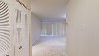 This single-floor residence offers a thoughtfully organized layout comprising a living room, bedroom, kitchen, bathroom, and closet. The living room is a generously proportioned space featuring wall-to-wall carpeting in a neutral beige tone and a large sliding glass door dressed with vertical blinds, allowing abundant natural light to fill the room. A ceiling-mounted light fixture adds to the bright, airy ambiance.

The bedroom is a well-sized private retreat, also carpeted and finished in soft, neutral tones, with a window fitted with horizontal blinds. It connects directly to a walk-in closet equipped with dual hanging rods on opposing walls, offering practical storage capacity.

The kitchen is efficiently designed with warm honey-toned wood cabinetry spanning both upper and lower sections, light countertops, and tile-style flooring. White appliances — including a gas range, over-the-range microwave, and dishwasher — complete the functional setup.

The bathroom features tile flooring, a full-size bathtub and shower combination with classic white square tile surround, a toilet, and a vanity with ample counter space. Clean white finishes throughout give the bathroom a crisp, tidy feel. Overall, the space presents a clean, neutral palette and a functional, comfortable layout.