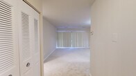 This single-floor residence offers a thoughtfully organized layout comprising a living room, bedroom, kitchen, bathroom, and closet. The living room is a generously proportioned space featuring wall-to-wall carpeting in a neutral beige tone and a large sliding glass door dressed with vertical blinds, allowing abundant natural light to fill the room. A ceiling-mounted light fixture adds to the bright, airy ambiance.

The bedroom is a well-sized private retreat, also carpeted and finished in soft, neutral tones, with a window fitted with horizontal blinds. It connects directly to a walk-in closet equipped with dual hanging rods on opposing walls, offering practical storage capacity.

The kitchen is efficiently designed with warm honey-toned wood cabinetry spanning both upper and lower sections, light countertops, and tile-style flooring. White appliances — including a gas range, over-the-range microwave, and dishwasher — complete the functional setup.

The bathroom features tile flooring, a full-size bathtub and shower combination with classic white square tile surround, a toilet, and a vanity with ample counter space. Clean white finishes throughout give the bathroom a crisp, tidy feel. Overall, the space presents a clean, neutral palette and a functional, comfortable layout.