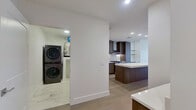 This thoughtfully designed one-bedroom, one-bathroom residence offers a refined modern aesthetic across a well-appointed single floor. The open-concept kitchen and living area serves as the heart of the home, showcasing rich dark walnut cabinetry that extends floor-to-ceiling, a striking marble-veined backsplash, stainless steel appliances, and a generous white quartz island that anchors the space. Light wood flooring flows throughout the main living areas, complementing the clean, minimalist design. Floor-to-ceiling windows frame sweeping high-rise views of the city skyline, filling the living area with abundant natural light and a dramatic urban backdrop. The spacious primary bedroom features plush carpeting, crisp white walls, and large windows offering continued panoramic city views, along with a convenient built-in floating desk nook. The bathroom is appointed with large-format marble-look porcelain tile, a glass-enclosed walk-in shower with a mosaic tile floor, a wall-mounted vanity with quartz countertop, and recessed lighting for a polished, spa-inspired atmosphere. A dedicated laundry room houses a stacked LG washer and dryer unit on marble-look tile flooring. The hallway connects each space efficiently, completing this well-organized and stylish urban residence.