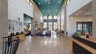This impressive single-floor lobby space makes an immediate statement with its soaring double-height ceilings, creating a grand and open atmosphere. The ceiling is finished in a bold teal hue, providing a striking visual contrast against the exposed brick columns and white shiplap wall panels that frame the space. Dramatic sputnik-style chandeliers hang at varying heights, serving as focal art pieces while casting warm, ambient light throughout.

The flooring is polished concrete, lending an industrial-chic aesthetic that complements the overall modern design sensibility. Natural light pours in through expansive windows and clerestory glazing, connecting the interior to the outdoors and enhancing the airy feel of the space.

The seating area is thoughtfully arranged with mid-century modern furnishings, including leather accent chairs, upholstered lounge pieces, and wooden occasional tables, creating intimate conversation zones within the larger open floor plan. A reception or service counter is visible to one side, indicating a welcoming, functional purpose for the space.

Decorative details such as stacked log accents and sculptural lighting elements add warmth and character. Overall, the space balances industrial materials with curated furnishings to deliver a sophisticated, welcoming, and visually dynamic environment.