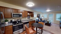 Located on Fifth Street SE in Puyallup, Washington, this single-floor unit offers a well-organized layout that balances functionality and comfort. The open-concept kitchen and dining area form the heart of the space, featuring warm medium-toned wood cabinetry, stainless steel appliances including a refrigerator, electric range, built-in microwave, and dishwasher, along with a double-basin sink and grey countertops. A compact round dining table with leather-upholstered chairs sits adjacent to the kitchen, providing a convenient everyday dining spot. Hardwood-style flooring runs throughout this area, adding warmth and visual continuity.

The living room flows naturally from the kitchen and dining space, featuring plush carpeting, a patio door, and windows fitted with vertical blinds that allow natural light to filter in. The bedroom is a calm and well-proportioned retreat with carpet flooring, a large window offering outdoor views, and ample wall space. The bathroom is equipped with a full tub and shower combination, hardwood-style flooring, and clean, neutral finishes. Throughout the unit, warm beige walls and consistent flush-mount ceiling lighting contribute to a cohesive, inviting atmosphere.