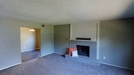 This two-floor home offers a practical and comfortable layout designed for everyday living. The living room serves as the heart of the main level, featuring a classic brick fireplace with a built-in mantle that anchors the space and provides a focal point for the room. Plush wall-to-wall carpeting in a neutral gray tone adds warmth underfoot, while the textured popcorn ceiling lends a traditional character to the space. Soft, neutral gray walls create a calm and versatile backdrop that complements a variety of décor styles. A doorway from the living room leads into an adjacent room, visible in the background and illuminated by a chandelier, suggesting a dining area or additional living space. The open sightlines between rooms enhance the sense of connectivity throughout the main floor. Consistent use of neutral tones and classic materials such as brick and carpet give the home a timeless, welcoming ambiance. With two floors of living space, the home provides ample room for both everyday activities and private retreat, making it a well-rounded residential property suited for a range of lifestyles.