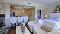 This single-floor space showcases a clean, contemporary design centered around an open-concept layout that connects the living area and kitchen seamlessly. The living room is furnished with crisp white upholstered seating, including a sofa and armchair accented with patterned throw pillows in warm orange and gray tones. A sleek white coffee table anchors the space, decorated with modern accent pieces that reinforce the unit's polished aesthetic.

The kitchen is defined by a peninsula-style breakfast bar topped with light-toned countertops and lined with tall white bar stools featuring cut-out back details. Pendant lighting and recessed fixtures illuminate the area, highlighting a stainless steel sink, dark appliances, and a decorative floral arrangement that adds a touch of warmth. A range hood is centrally positioned above the cooking area, contributing to the kitchen's functional yet stylish character.

Dark hardwood flooring runs throughout, providing a striking contrast against the predominantly white furnishings and neutral walls. A framed artwork piece adorns one wall, while an open doorway hints at additional rooms beyond, including what appears to be a closet or bedroom. The overall ambiance is bright, airy, and modern.