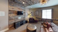 This well-appointed single-floor communal space masterfully combines rustic charm with contemporary design. The layout flows between several distinct areas, each thoughtfully curated with cohesive furnishings and décor. A reception or business area features a dark counter with bar-height seating, pendant lighting, and bright windows that allow natural light to pour in — ideal for administrative or concierge functions. Adjacent to it, a seating nook offers black leather booth-style banquettes with a compact table and a mounted screen, creating a functional yet comfortable spot for work or casual meetings. The lounge and game room serve as the centerpiece of the space, anchored by a striking reclaimed wood accent wall that extends across both the wall and ceiling, lending warmth and texture to the room. A wall-mounted flat-screen television, a media console, and plush seating — including deep purple armchairs and a leather ottoman — create an inviting entertainment zone. A shuffleboard table adds a recreational element, perfect for socializing. Framed artwork adorns the neutral-toned walls throughout, while a statement sputnik chandelier and floor lamps provide layered lighting. Carpet tile flooring ties the areas together for a cohesive, modern-rustic aesthetic.