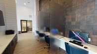 Located in Richmond, Texas, this contemporary single-story community clubhouse on Berkeley Knoll Circle is thoughtfully designed to serve as a versatile social and professional hub. The space is defined by its dramatic soaring ceilings and expansive floor-to-ceiling windows framed in sleek black that flood the interiors with natural light while offering views of the surrounding landscaped grounds and community buildings.

The open-plan layout features distinct zones that transition seamlessly from one to the next. A generously proportioned lounge area anchors the space, furnished with neutral-toned sofas, wooden accent chairs, and a layered area rug, all set against shiplap-clad white columns and warm wood-look tile flooring in a herringbone pattern. A bar-height counter with a white marble surface and blue velvet stools sits before a striking wall of glossy dark tile, complete with a mounted television and pendant lighting above.

The workspace area includes individual computer stations separated by frosted privacy panels alongside an accent wall featuring rich wood-toned tile. Semi-private high-back booth seating with integrated power outlets provides additional flexible work areas, complemented by a glass-enclosed private office. The overall ambiance balances modern sophistication with warmth, making the space equally suited for work, relaxation, and community gatherings.