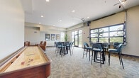 This single-story community clubhouse is thoughtfully designed to offer a variety of social and recreational spaces within a cohesive, modern layout. The main hall serves as the central artery of the facility, showcasing soaring ceilings, warm track lighting, light wood-look flooring, and black-framed glass partition walls that provide visual connectivity between areas while maintaining defined zones.

The primary gathering area is generously proportioned, featuring patterned carpet flooring and multiple high-top tables paired with teal upholstered bar-height chairs. A wall-mounted flat-screen television anchors one end of the room, while large windows and a full-width overhead garage-style door along one wall allow abundant natural light and views of the surrounding greenery, creating an indoor-outdoor feel when open.

A standout feature is the full-length shuffleboard table positioned along one side of the room, complemented by additional game tables nearby. Framed artwork and potted plants add character throughout the space.

Tucked adjacent to the main area is a dedicated media lounge, furnished with plush leather armchairs, side tables, and a wall-mounted television set within a tray-ceiling alcove — offering a more intimate retreat. Recessed lighting throughout the facility ensures consistent illumination across all zones.