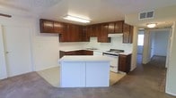 This single-floor space is centered around a functional and well-appointed kitchen that serves as the heart of the layout. The kitchen features rich, dark wood cabinetry that extends along two walls in an L-shaped configuration, providing generous upper and lower storage. A white central island adds additional counter space and acts as a natural dividing element between the kitchen and the adjacent carpeted living area. White appliances, including a gas range and an under-counter dishwasher, are neatly integrated into the design, complementing the light-colored countertops. A stainless steel sink is positioned beneath the upper cabinetry, and a range hood is mounted above the cooktop for ventilation. Overhead, a flush-mounted fluorescent light fixture provides bright, even illumination throughout the kitchen workspace. The textured ceiling adds a subtle classic character to the interior. The transition from the tiled kitchen floor to the neutral wall-to-wall carpet in the adjoining area creates a clear delineation between functional and living spaces. Overall, the space presents a straightforward, practical design that prioritizes utility and ease of everyday living.