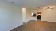 This single-floor space offers an open and functional layout that seamlessly connects the living area to the kitchen. The living area is bright and airy, finished with warm hardwood-style flooring that extends throughout the space, complemented by clean white walls and white baseboards that lend a fresh, neutral atmosphere. A textured ceiling adds a subtle design detail overhead, while recessed vents ensure efficient climate control.

The kitchen is positioned at the rear of the open floor plan and is defined by rich, warm-toned wood cabinetry that provides ample storage. Black appliances, including a refrigerator and a range with overhead ventilation, give the kitchen a bold, classic contrast against the cabinetry. A peninsula-style counter serves as a natural divider between the kitchen and living areas, offering additional prep space and a casual seating opportunity.

A closet with bi-fold doors is conveniently situated near the living area, providing practical storage. A secondary door leads to an adjacent room or exterior access, adding to the overall connectivity of the layout. The space is efficiently organized, making it easy to navigate while maintaining an open, welcoming feel throughout.