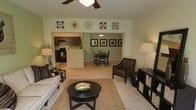 This single-story home features an open-concept layout that seamlessly connects the living room, dining area, and kitchen into one cohesive living space. The living room is finished with light-toned carpet and pale green walls, creating a calm, neutral backdrop. A ceiling fan with a light fixture anchors the room from above, while a sofa, armchair, round dark-wood coffee table, and woven area rug provide a comfortable and well-appointed arrangement. Decorative wall art in a coordinated color palette adds visual interest above the seating area. A pass-through opening connects the living space to the kitchen, which features dark cabinetry, black appliances, and a white countertop peninsula that also serves as an informal divider. Adjacent to the kitchen, the dining area accommodates a dark wood table and chair set beneath a pendant light fixture, with a series of framed artwork arranged neatly on the wall. A storage unit with open shelving sits at the edge of the living room, offering both function and display space. The overall ambiance is bright, tidy, and practical.