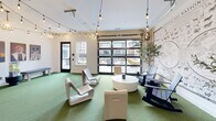 Located at 25 W Seventh Street in Richmond, Virginia, this thoughtfully designed dog-friendly amenity space delivers a welcoming and whimsical atmosphere for pets and their owners alike. The single-floor layout makes clever use of its cozy footprint, offering a bright and open lounge area anchored by lush green turf flooring that sets a playful, park-like tone indoors. String lights draped across the ceiling cast a warm, inviting glow, complemented by globe pendant fixtures that add a decorative touch. A striking "Dogs of All Nations" world map mural serves as a bold focal point on the feature wall, accented by a vibrant teal accent wall alongside it. Modern sculpted white chairs, Adirondack seating, and a round coffee table furnish the lounge area, while decorative potted trees add a natural element. On an adjacent wall, a gallery of regal, Renaissance-style dog portrait canvases injects personality and charm, with a tennis ball dispenser serving as a fun, functional accent. A large glass panel garage-style door floods the space with natural light and connects it visually to the outdoors. A separate grooming or washing area features floor-to-ceiling tiled walls adorned with dog silhouette patterns in navy and white, polished concrete floors, and warm brass wall sconces, combining both style and utility seamlessly.