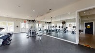 This fitness center offers a comprehensive workout environment designed to accommodate a wide range of exercise routines. The space is laid out across a single floor with an open, functional floor plan that maximizes usability and traffic flow. Durable dark rubber flooring spans the entire area, providing traction and impact absorption suitable for high-intensity activity.

The cardio zone is well-stocked with multiple treadmills, elliptical trainers, and upright bikes, all positioned along walls lined with large windows that allow generous natural light to fill the room and offer pleasant outdoor views of surrounding greenery. A wall-mounted flat-screen TV is positioned above the cardio equipment for entertainment during workouts.

A dedicated strength training area features a dual cable cross machine alongside a weight bench, enabling a variety of resistance exercises. Floor-to-ceiling mirrors line one wall, visually expanding the space and allowing users to monitor their form. A stability ball rack stocked with multiple sizes of exercise balls rounds out the functional accessories available.

Recessed lighting throughout the ceiling ensures consistent illumination across all zones. The neutral color palette of white walls and light trim creates a clean, bright atmosphere. Signage displaying fitness center rules is posted on the wall, and a sanitizing dispenser is mounted near the entrance for hygiene convenience.