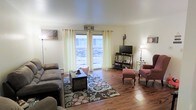 This single-floor space offers a comfortable and well-appointed living environment. The living room serves as the centerpiece of the home, featuring rich hardwood flooring that extends throughout and lends a warm, grounded aesthetic to the space. Large windows dressed with light, sheer cream curtains allow generous amounts of natural light to fill the room, creating a bright and airy ambiance. A sliding glass door provides a view of an exterior stone feature, further connecting the interior to the outdoors. The living room is thoughtfully arranged with ample seating, including a plush sofa and a classic accent chair, centered around a decorative area rug with a floral motif. A media console with an open shelving unit houses a flat-screen television and organized media storage, making the room equally suited for relaxation and entertainment. A wall-mounted air conditioning unit ensures year-round comfort. Soft lamp lighting complements the natural light, adding to the room's cozy and welcoming character. The clean, neutral wall tones throughout provide a versatile backdrop, allowing the furnishings and natural elements to stand out. Overall, this space presents a functional and inviting single-level layout ideal for everyday living.