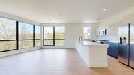 This contemporary single-floor residence showcases a thoughtfully designed open-concept layout with clean lines and a bright, neutral color palette throughout. The expansive living and kitchen area is anchored by large black-framed windows that flood the space with natural light and offer treetop views, creating a seamless connection to the outdoors. Light wood-tone flooring runs throughout, lending warmth and continuity to every room.

The kitchen is a standout feature, centered around a generous white waterfall island with an undermount sink and matte black faucet. Upper cabinetry in soft white tones pairs with navy blue lower cabinets for a bold two-tone contrast. Stainless steel appliances, including a French door refrigerator and built-in microwave, complete the modern aesthetic.

The bedrooms are well-proportioned, each featuring the same light hardwood flooring, large black-framed windows with leafy outdoor views, and access to closet space. The living area extends to a private balcony through sliding glass doors, offering an outdoor retreat with tree-lined scenery.

The bathroom is finished with striking marble-look tile on both the floors and shower walls, complemented by a sleek floating vanity in charcoal grey with gold hardware, a round gold-framed mirror, and matte black fixtures — delivering a refined, spa-like atmosphere.