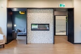 Located at 1425 Block Line Rd in Kitchener, Ontario, this well-appointed amenity space offers a thoughtfully designed, open-concept layout across a single floor. Light-toned hardwood-style flooring flows throughout, creating a cohesive and welcoming atmosphere.

The main gathering area serves as the heart of the space, combining a dining zone and lounge seating in one generous, light-filled room. Large windows along the perimeter flood the interior with natural light and frame views of surrounding greenery. A sleek, wall-mounted linear fireplace set against a mosaic tile feature wall adds warmth and visual interest, while a tray ceiling detail with recessed lighting and a contemporary geometric pendant fixture lends architectural sophistication.

A full kitchenette is seamlessly integrated into the main area, featuring stainless steel appliances — including a full-sized refrigerator, electric range, and dishwasher — complemented by white cabinetry and a decorative mosaic tile backsplash. Direct access to an outdoor space is provided through a glass door, enhancing connectivity with the outdoors.

A separate, more intimate lounge room offers a quieter retreat, finished with warm wood panel walls, a natural fiber area rug, a leather sofa, and blue accent chairs — ideal for private conversations or relaxed gatherings. A connecting hallway links the areas efficiently, completing this versatile and elegantly appointed communal space.