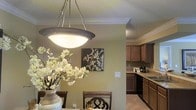 This single-floor residence offers a thoughtfully designed interior that blends functionality with classic elegance. The dining area serves as a welcoming focal point, highlighted by a striking bowl-shaped pendant light suspended from the ceiling, casting a warm, inviting glow over the dining table and chairs below. Crown molding runs along the ceiling perimeter, adding a refined architectural detail that elevates the overall aesthetic of the space.

The warm, neutral wall tones create a cohesive and calming atmosphere that flows seamlessly from the dining area into the adjacent kitchen. The kitchen is well-equipped, featuring rich, dark wood cabinetry that provides ample storage, dark countertops, and tile flooring that is both practical and stylish. The layout allows for an easy transition between meal preparation and dining, making the space ideal for both everyday living and entertaining.

Decorative touches, including a large floral arrangement in a woven vase and tasteful wall art, add warmth and personality to the interior. The textured ceiling detail in the kitchen area provides a subtle contrast to the smooth, molded ceilings in the dining space. Overall, this home presents a harmonious blend of classic architectural details and comfortable, functional design.