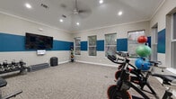 This single-room fitness studio offers a well-equipped workout environment within a compact, efficiently designed footprint. The space features vaulted ceilings accented with crown molding, recessed lighting, and a ceiling fan that enhances air circulation during workouts. A bold two-tone color scheme — warm beige on the lower walls and a vibrant blue horizontal band — gives the studio a dynamic, energetic aesthetic.

Multiple windows along two walls allow natural light to fill the space while providing outdoor views of surrounding greenery. The neutral, textured carpet flooring provides a comfortable surface suitable for a variety of exercises.

Equipment is thoughtfully arranged throughout the room. A row of spin bikes is positioned along one side, ideal for cycling classes or individual cardio sessions. A rack of kettlebells and step platforms supports strength and functional training, while a colorful medicine ball rack adds versatility for conditioning workouts. A large wall-mounted flat-screen television serves as a focal point, complemented by a whiteboard for class schedules or workout displays. A foam roller and additional accessories are mounted near the entry door, completing the studio's well-rounded fitness offerings.