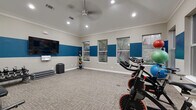 This single-room fitness studio offers a well-equipped workout environment within a compact, efficiently designed footprint. The space features vaulted ceilings accented with crown molding, recessed lighting, and a ceiling fan that enhances air circulation during workouts. A bold two-tone color scheme — warm beige on the lower walls and a vibrant blue horizontal band — gives the studio a dynamic, energetic aesthetic.

Multiple windows along two walls allow natural light to fill the space while providing outdoor views of surrounding greenery. The neutral, textured carpet flooring provides a comfortable surface suitable for a variety of exercises.

Equipment is thoughtfully arranged throughout the room. A row of spin bikes is positioned along one side, ideal for cycling classes or individual cardio sessions. A rack of kettlebells and step platforms supports strength and functional training, while a colorful medicine ball rack adds versatility for conditioning workouts. A large wall-mounted flat-screen television serves as a focal point, complemented by a whiteboard for class schedules or workout displays. A foam roller and additional accessories are mounted near the entry door, completing the studio's well-rounded fitness offerings.