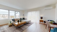 This well-designed single-floor unit offers a clean, contemporary aesthetic throughout, characterized by crisp white walls, light wood-style flooring, and an abundance of natural light. The spacious living room serves as the heart of the unit, featuring large windows and a sliding glass door fitted with vertical blinds that open to an outdoor balcony, filling the space with daylight and offering views of the surrounding neighborhood. The open layout flows seamlessly into a dedicated dining area, which includes a wall-mounted mini-split climate control unit and its own access to the balcony, creating a bright and airy atmosphere ideal for everyday living. The galley-style kitchen is efficiently organized with high-gloss white cabinetry, dark granite countertops, and stainless steel appliances including a range, microwave, and refrigerator, all complemented by recessed lighting and light hardwood flooring. The bedroom is a serene retreat, featuring plush carpeting, geometric wall art, a built-in closet with sliding wood-panel doors, and a mini-split unit for personalized comfort. The full bathroom is finished with wood-tile flooring and includes a shower-tub combination. Throughout the unit, the consistent use of light tones and modern finishes creates a cohesive and inviting living environment.