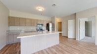 This single-floor residence is designed with a clean, contemporary aesthetic that emphasizes both functionality and comfort. The heart of the home is the open-concept kitchen, which flows seamlessly into the living area. The kitchen is outfitted with a full suite of stainless steel appliances, including a refrigerator, range, and over-the-range microwave. Warm-toned, shaker-style cabinetry lines the walls, complemented by granite-look countertops that extend to a generously sized kitchen island with an integrated sink. This island serves as both a prep space and a natural gathering point, adding to the open and airy feel of the area. Light wood-look flooring runs throughout the main living spaces, lending warmth and cohesion to the design.

A hallway connects the main living area to the private spaces of the home. The bedroom features plush carpeting underfoot and is equipped with a double-door closet offering built-in shelving and hanging rods for ample storage. The bathroom is well-appointed, featuring a granite-look vanity countertop with a single-basin sink, a large mirror, and a full tub-shower combination. Neutral wall tones and consistent finishes throughout the space create a cohesive and welcoming atmosphere.