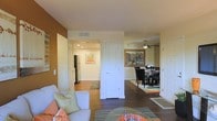 This single-floor residence offers a thoughtfully designed open-concept layout that seamlessly connects the living, dining, and kitchen areas. The living room serves as a welcoming focal point, featuring a comfortable sofa accented with decorative pillows in warm orange and green tones, set atop a patterned area rug. A rich, earthy accent wall adds depth and warmth to the space, complementing the neutral ceiling and trim throughout.

Hardwood-style flooring extends through the main living and dining areas, lending a cohesive and polished look. The dining area is furnished with a dark-stained table and matching chairs, topped by a ceiling fan with integrated lighting — both functional and stylish. A framed cityscape artwork adorns the dining room wall, adding a contemporary artistic touch.

The kitchen is accessible via an open corridor and features dark appliances, including a refrigerator, with light-toned cabinetry and recessed lighting overhead. A hallway with a small area rug leads from the kitchen, suggesting additional rooms beyond. The overall ambiance is warm, inviting, and well-lit, with recessed lighting and natural tones creating a comfortable and functional living environment throughout the home.
