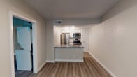 This single-floor unit offers a clean, modern layout designed for comfortable everyday living. The open-concept living and kitchen area serves as the heart of the space, where light hardwood-style flooring runs continuously underfoot, creating a cohesive and airy feel throughout. The kitchen is outfitted with a full suite of stainless steel appliances, including a refrigerator, electric range, and over-the-range microwave. White shaker-style cabinetry provides ample storage, while the peninsula countertop with a stainless steel sink offers both workspace and a natural divide between the kitchen and living area. Track lighting illuminates the kitchen, and a textured ceiling adds subtle character overhead. The bedroom is a well-proportioned space featuring the same hardwood-style flooring, a ceiling fan with integrated lighting, vaulted ceiling detail, a window with blinds that brings in natural light, and a sliding door closet. An additional room or den is accessible directly from the bedroom, offering flexible use as a home office or secondary space. The bathroom features a white vanity with a gray countertop, an undermount sink, a framed mirror with overhead vanity lighting, and a full tub-shower combination. Neutral tones throughout the unit give it a fresh, move-in ready appearance.