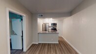 This single-floor unit offers a clean, modern layout designed for comfortable everyday living. The open-concept living and kitchen area serves as the heart of the space, where light hardwood-style flooring runs continuously underfoot, creating a cohesive and airy feel throughout. The kitchen is outfitted with a full suite of stainless steel appliances, including a refrigerator, electric range, and over-the-range microwave. White shaker-style cabinetry provides ample storage, while the peninsula countertop with a stainless steel sink offers both workspace and a natural divide between the kitchen and living area. Track lighting illuminates the kitchen, and a textured ceiling adds subtle character overhead. The bedroom is a well-proportioned space featuring the same hardwood-style flooring, a ceiling fan with integrated lighting, vaulted ceiling detail, a window with blinds that brings in natural light, and a sliding door closet. An additional room or den is accessible directly from the bedroom, offering flexible use as a home office or secondary space. The bathroom features a white vanity with a gray countertop, an undermount sink, a framed mirror with overhead vanity lighting, and a full tub-shower combination. Neutral tones throughout the unit give it a fresh, move-in ready appearance.