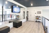 Located at 3050 Circle 75 Pkwy SE in Atlanta, Georgia, this fitness studio offers a well-appointed, single-floor workout space designed to accommodate a variety of training modalities. The interior is characterized by a striking dark ceiling contrasted against light gray walls, creating a sleek, contemporary aesthetic. Warm-toned wood-look flooring runs throughout, lending an approachable feel to the otherwise bold design.

The space is divided into distinct functional zones. One area is dedicated to functional and resistance training, featuring wall-mounted TRX suspension straps, resistance bands, and a large flat-screen display for guided workouts. Full-length mirrors along the walls enhance the sense of openness and allow for form monitoring during exercise.

An adjacent zone accommodates low-impact and flexibility training, housing a pilates reformer, a BOSU balance trainer, and a tall rack stocked with stability balls in assorted sizes. A dark foam mat panel is mounted to the wall nearby. The space is open along one side, with a sleek black cable-and-post railing separating the studio from an adjoining fitness area below, adding an architectural element while maintaining visual connectivity between zones. Recessed lighting throughout ensures the space is evenly and well-lit.