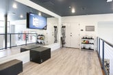 Located at 3050 Circle 75 Pkwy SE in Atlanta, Georgia, this fitness studio offers a well-appointed, single-floor workout space designed to accommodate a variety of training modalities. The interior is characterized by a striking dark ceiling contrasted against light gray walls, creating a sleek, contemporary aesthetic. Warm-toned wood-look flooring runs throughout, lending an approachable feel to the otherwise bold design.

The space is divided into distinct functional zones. One area is dedicated to functional and resistance training, featuring wall-mounted TRX suspension straps, resistance bands, and a large flat-screen display for guided workouts. Full-length mirrors along the walls enhance the sense of openness and allow for form monitoring during exercise.

An adjacent zone accommodates low-impact and flexibility training, housing a pilates reformer, a BOSU balance trainer, and a tall rack stocked with stability balls in assorted sizes. A dark foam mat panel is mounted to the wall nearby. The space is open along one side, with a sleek black cable-and-post railing separating the studio from an adjoining fitness area below, adding an architectural element while maintaining visual connectivity between zones. Recessed lighting throughout ensures the space is evenly and well-lit.