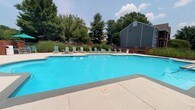 Barclay offers a well-appointed outdoor amenity space centered around a generously sized swimming pool with crystal-clear blue water. The pool deck is finished in smooth concrete, providing ample space for movement and leisure around the water's edge. A row of light green lounge chairs lines one side of the pool, offering residents comfortable spots to relax and soak up the sun. Decorative brick pillars and ornamental grasses add a touch of structured landscaping to the perimeter, complementing the natural surroundings. Mature trees border the area, providing a lush green backdrop that enhances the sense of privacy and tranquility. A residential building with a dark-colored gabled roof is visible in the background, contributing to the community feel of the setting. The open sky above allows for abundant natural light throughout the day, making this outdoor space bright and inviting. Overall, the pool area at Barclay is thoughtfully designed to serve as a central gathering and recreation hub, combining functional amenities with attractive landscaping for a comfortable and enjoyable outdoor experience.