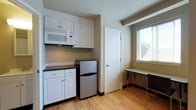 This single-floor unit offers a straightforward, functional layout designed for comfortable everyday living. The sleeping area is finished with warm light hardwood flooring that runs throughout the space, neutral painted walls, and a window with blinds that allows natural light to fill the room. A twin bed frame with a simple wooden design anchors the space, and a dedicated desk and chair provide a practical area for work or study.

The kitchenette is efficiently arranged with white cabinetry offering ample storage, a dark countertop, an over-the-counter microwave, and a compact stainless steel refrigerator. A window positioned near the dining area brings in additional natural light, and a small table with chairs creates a convenient spot for meals.

The private bathroom features a clean, minimal design with light-toned flooring, a white vanity with under-cabinet storage, a mirrored medicine cabinet, and a well-lit overhead fixture that brightens the space. The overall ambiance of the unit is practical and uncluttered, with a consistent neutral color palette and durable finishes that lend a tidy, move-in-ready feel throughout every area of the space.