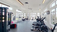 Located at 3400 Kyle Crossing in Kyle, Texas, this single-story fitness center is designed to provide a bright and motivating workout environment. The space is characterized by soaring ceilings with large, multi-pane windows lining the walls, flooding the interior with an abundance of natural light and offering views of the surrounding community. Modern pendant and linear light fixtures complement the natural illumination, while ceiling fans promote airflow throughout the open floor plan.

The fitness center is laid out to maximize usability across its spacious interior. A dedicated cardio zone features a row of treadmills, ellipticals, stationary bikes, and stair climbers, all equipped with individual display screens. A free weights area offers a full dumbbell rack alongside adjustable benches and large wall mirrors, allowing users to monitor their form during strength training sessions.

A functional training corner houses a multi-station cable and resistance rig complete with TRX suspension trainers, medicine balls, kettlebells, resistance bands, and a plyo box — supporting a wide variety of functional and cross-training workouts. The flooring throughout consists of durable, cushioned gray athletic material suited for high-traffic use. A water fountain station is conveniently located near the entrance. The clean, white walls and open layout create a welcoming and energetic ambiance suited for all fitness levels.