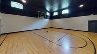 Located at 1717 E Belt Line Rd in Coppell, Texas, this single-floor indoor basketball facility is purpose-built for athletic activity and recreational use. The court is anchored by a polished hardwood floor finished in a warm natural tone, complete with clearly painted regulation court markings including a three-point arc, lane lines, and key areas. A professional-grade glass backboard with an orange rim and net is wall-mounted on one end, providing an authentic playing experience. The soaring ceiling height creates a true full-play environment, painted in a deep black that contrasts sharply with the bright white lower walls, lending the space a bold, modern aesthetic. A row of arched transom windows near the ceiling allows natural light to filter in, complementing the recessed and pendant overhead lighting that ensures consistent illumination throughout the court. The perimeter features dark baseboards that neatly frame the playing surface. Along one side wall, a set of glass-paneled double doors provides access to an adjacent mirrored fitness or stretching area, expanding the overall utility of the facility. The space is well-ventilated with visible HVAC vents in the ceiling, and safety exits are clearly marked. Overall, this facility offers a clean, well-appointed environment suited for both competitive play and casual recreation.