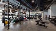 Located at 3615 Tryclan Drive in Charlotte, NC, this fitness center offers a generously proportioned, single-floor workout facility designed with a sleek industrial-modern aesthetic. The space features exposed ductwork and structural elements overhead, complemented by dark-painted ceilings and recessed track lighting that create a focused, energizing atmosphere. Large floor-to-ceiling windows along the exterior walls flood the space with natural light while offering views of the surrounding outdoor area.

The facility is thoughtfully divided into distinct training zones. A dedicated strength area houses multi-station power racks equipped with barbells, medicine balls, battle ropes, and an extensive array of free weights including a full rack of dumbbells, weight plates, and foam rollers. Adjustable benches provide versatility for a wide range of exercises. The cardio zone is lined with premium TRUE Fitness equipment, including stationary bikes, ellipticals, and treadmills, as well as a HIIT curved treadmill. A large mirrored wall with elegant draping accents enhances the cardio area's open feel. The durable rubberized flooring throughout ensures safety and comfort. Glass partitions and sliding doors connect additional adjoining areas, adding to the facility's functional flow and modern design.