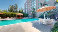 Located along the iconic Wilshire Boulevard in Los Angeles, this outdoor pool area offers a well-designed recreational retreat set against the backdrop of a multi-story building. The centerpiece is a generously sized rectangular swimming pool with vivid turquoise water, framed by a broad, smooth concrete deck that provides ample space for movement and relaxation.

Surrounding the pool, a curated selection of lounge chairs and seating arrangements are positioned to take advantage of the Southern California sunshine. A standout feature is the covered gazebo, outfitted with warm-toned striped curtains and comfortable seating beneath — an ideal shaded refuge on sunny days. Multiple market umbrellas in coordinating orange and white striped patterns are distributed throughout the deck, offering additional shade over white outdoor dining tables and chairs.

The space is thoughtfully landscaped with tall tropical palms, large-leafed banana plants, and manicured hedgerows that provide a sense of privacy and a lush, resort-like atmosphere. White fencing encloses a portion of the area, adding structure while maintaining an open, airy feel. Outdoor showers and clear safety signage reflect a commitment to functionality. The overall ambiance is bright, inviting, and characteristic of a classic Los Angeles outdoor amenity space.