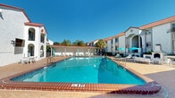 This three-floor property showcases a distinctive Spanish-Mediterranean architectural style, characterized by white stucco exteriors, red clay tile roofing, decorative arched entryways, and juliet balconies. The buildings are arranged around a central outdoor courtyard that serves as the heart of the property.

At the center of the outdoor space is a generously sized swimming pool with brick-tiled coping, offering varying depths clearly marked throughout. The surrounding pool deck is finished in warm-toned pavers, providing ample space for lounging. Comfortable outdoor seating is arranged across the deck, complemented by teal-accented umbrellas that add a pop of color against the bright Florida sky.

A dedicated poolhouse structure sits alongside the pool, featuring its own red tile roof, light blue entry doors, and an open covered porch lined with lounge chairs — ideal for shaded relaxation. On the opposite side of the pool deck, a built-in outdoor fireplace with a seating area creates an inviting gathering space for cooler evenings.

Mature palm trees are scattered throughout the property, enhancing the resort-like atmosphere and providing natural shade. The landscaping, open sightlines, and sun-drenched courtyard collectively contribute to a bright, welcoming ambiance. The multi-floor building structure overlooks the pool area, with balconies offering elevated views of the outdoor amenities.